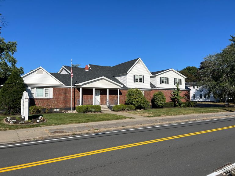 Two-story yellow building with green trim, arched windows, and a black paved driveway on a sunny day.