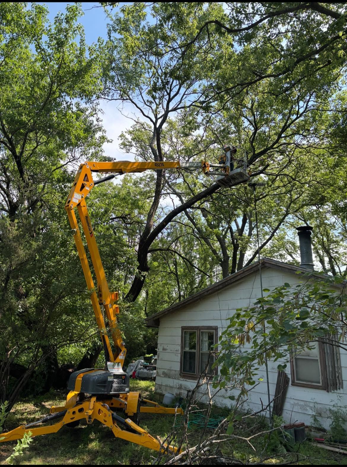 Large orange tree trimmer on a boom lift working beside a white house surrounded by trees