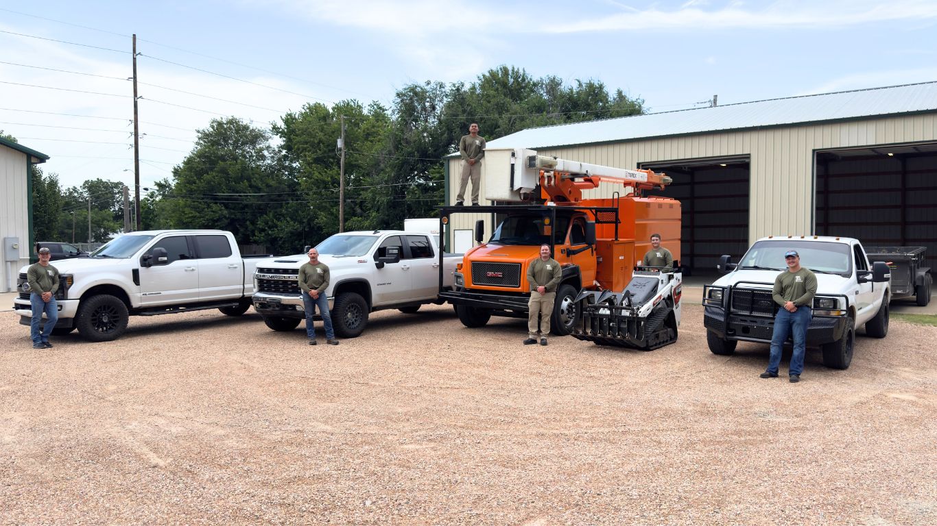 Utility trucks and workers lined up on a gravel lot beside a warehouse, with an orange machine in the center