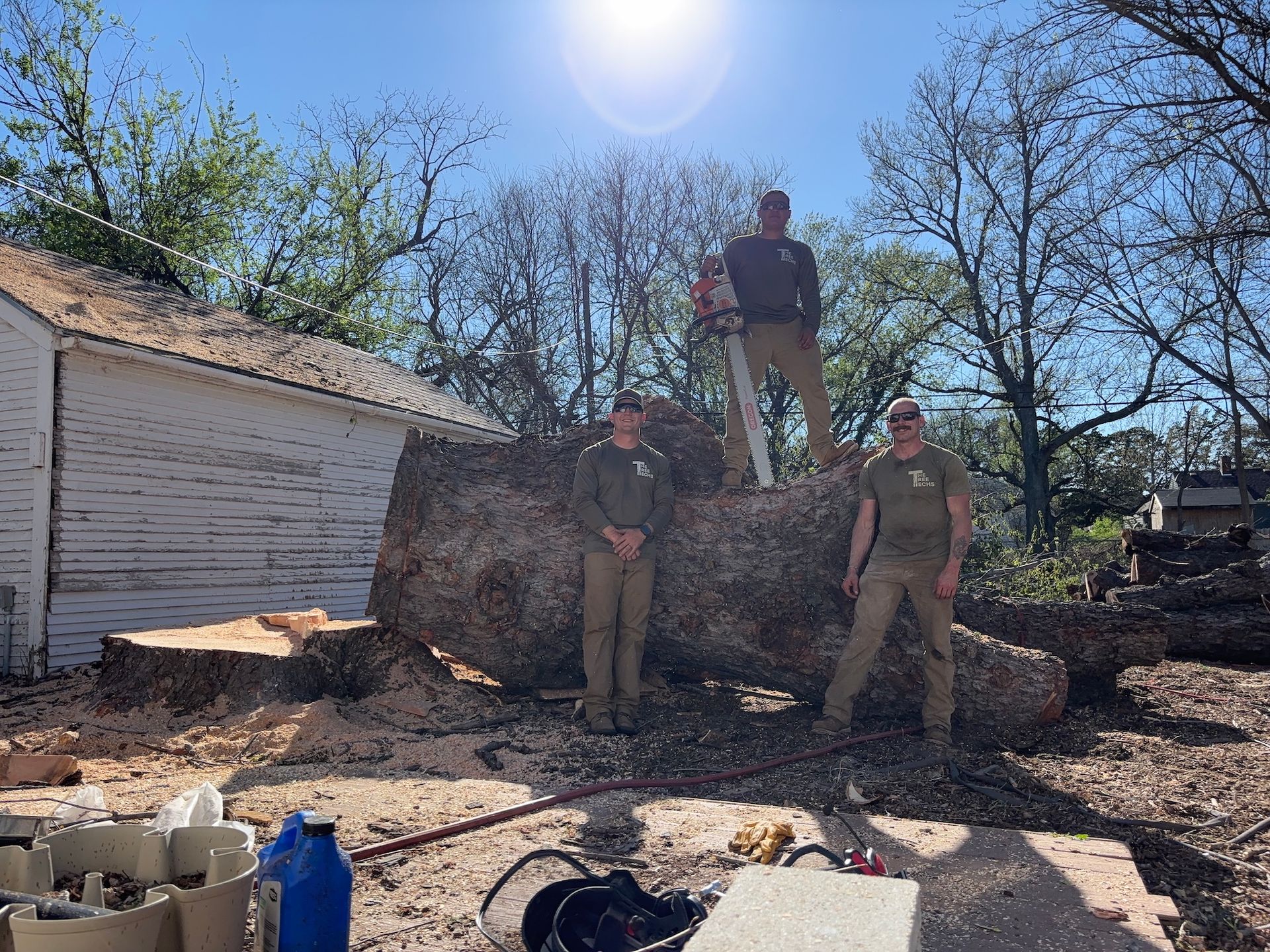 Three people in tan uniforms stand on a large tree trunk beside a white shed in a sunny yard.