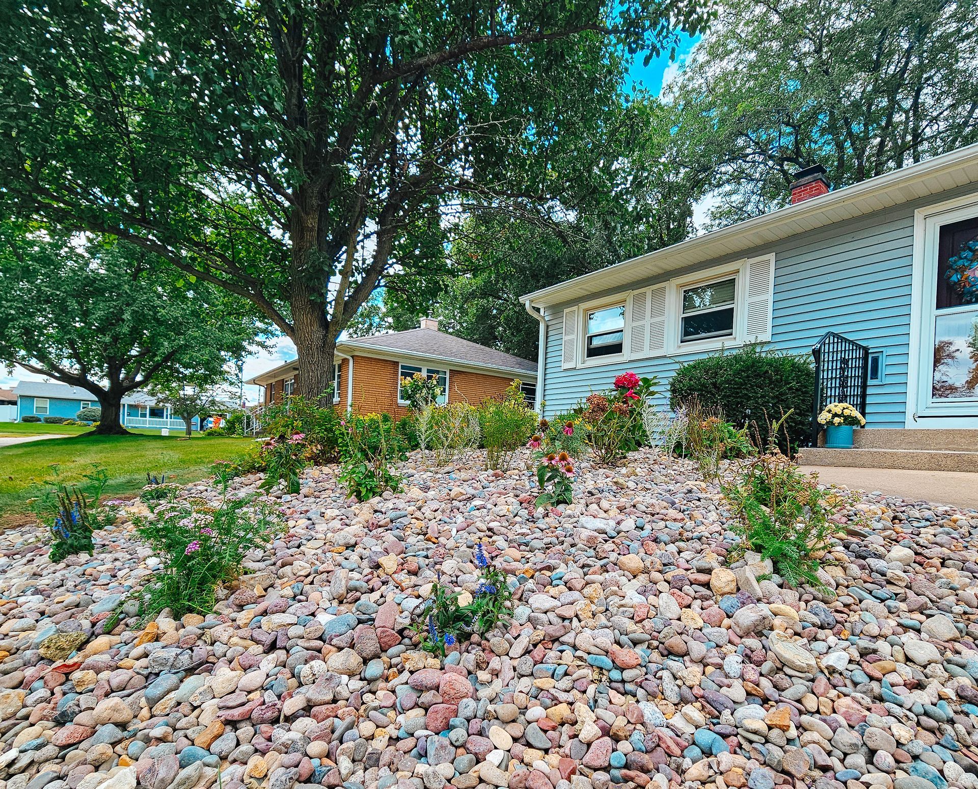 Front yard with colorful river rocks, a large tree, and a light blue house with a covered porch