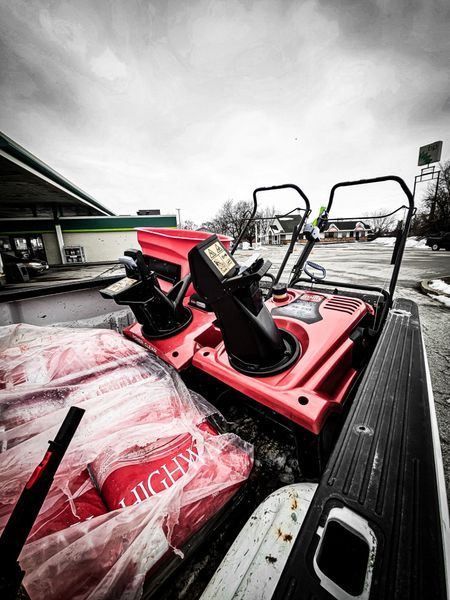 Two red snow blowers and bags of salt sit in the back of a truck in a parking lot on an overcast day.