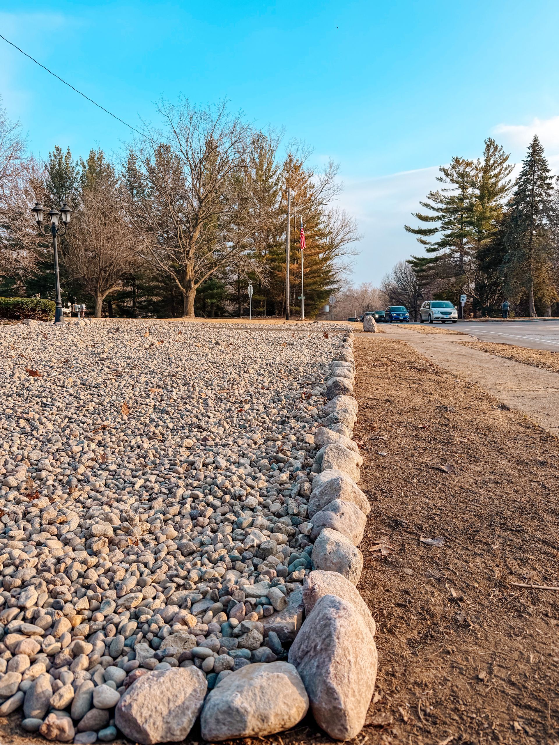 Rock-lined roadside beside a dirt path, with trees and a blue sky in the background.