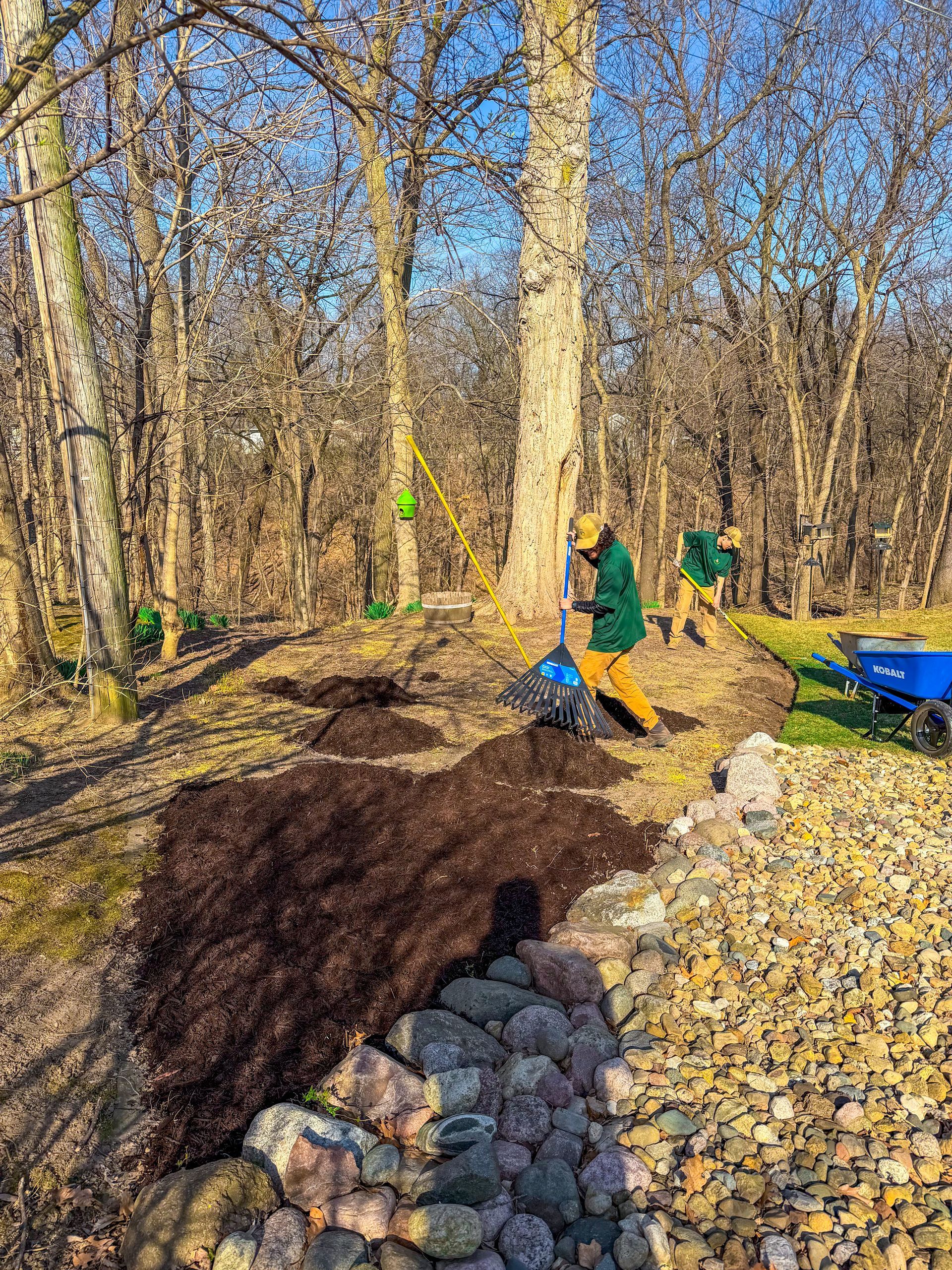 People working in a wooded area beside a gravel path and large tree trunk, with wheelbarrows and soil.