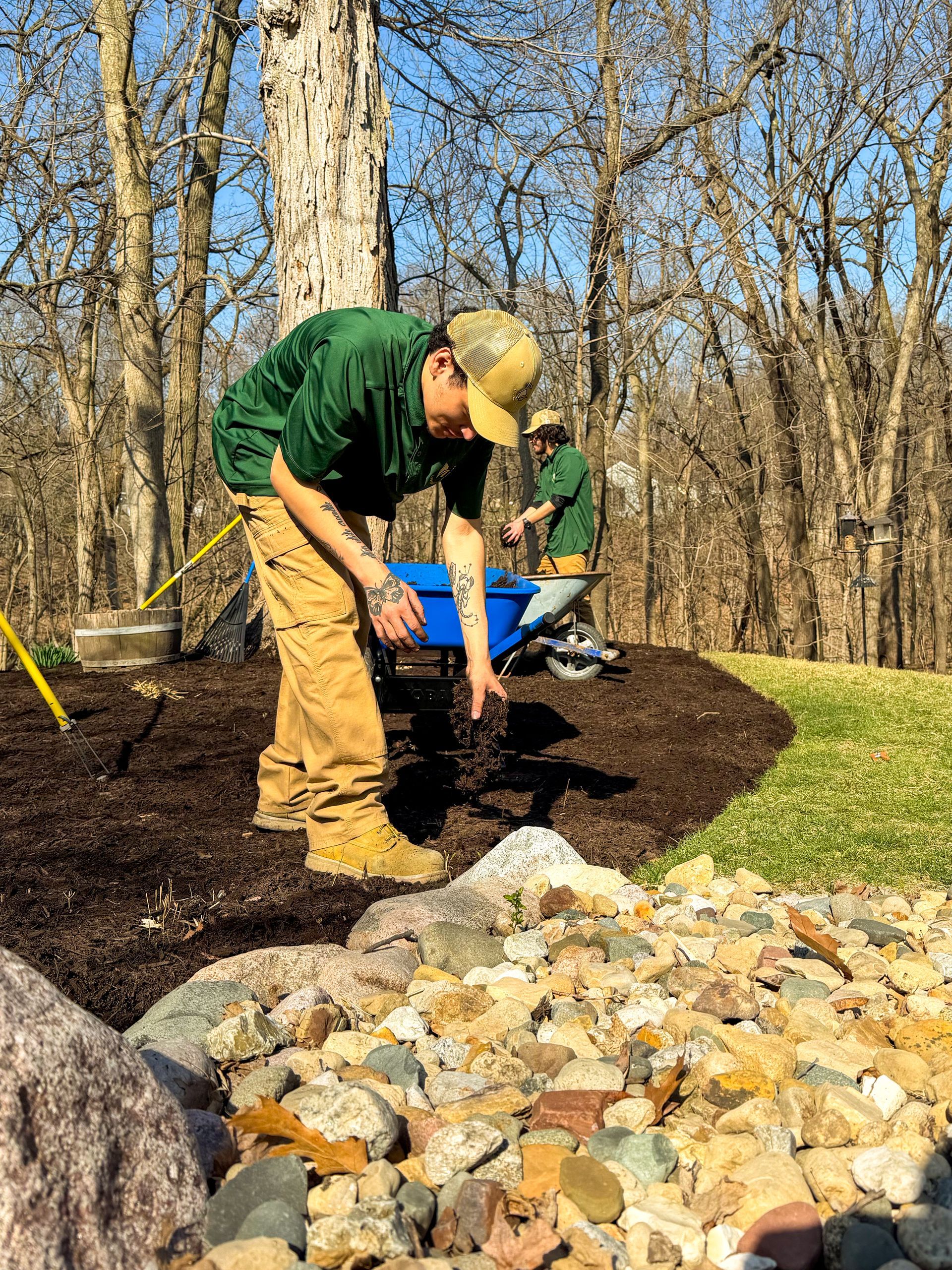 Person shoveling mulch beside a rock border in a wooded yard, with another person behind.