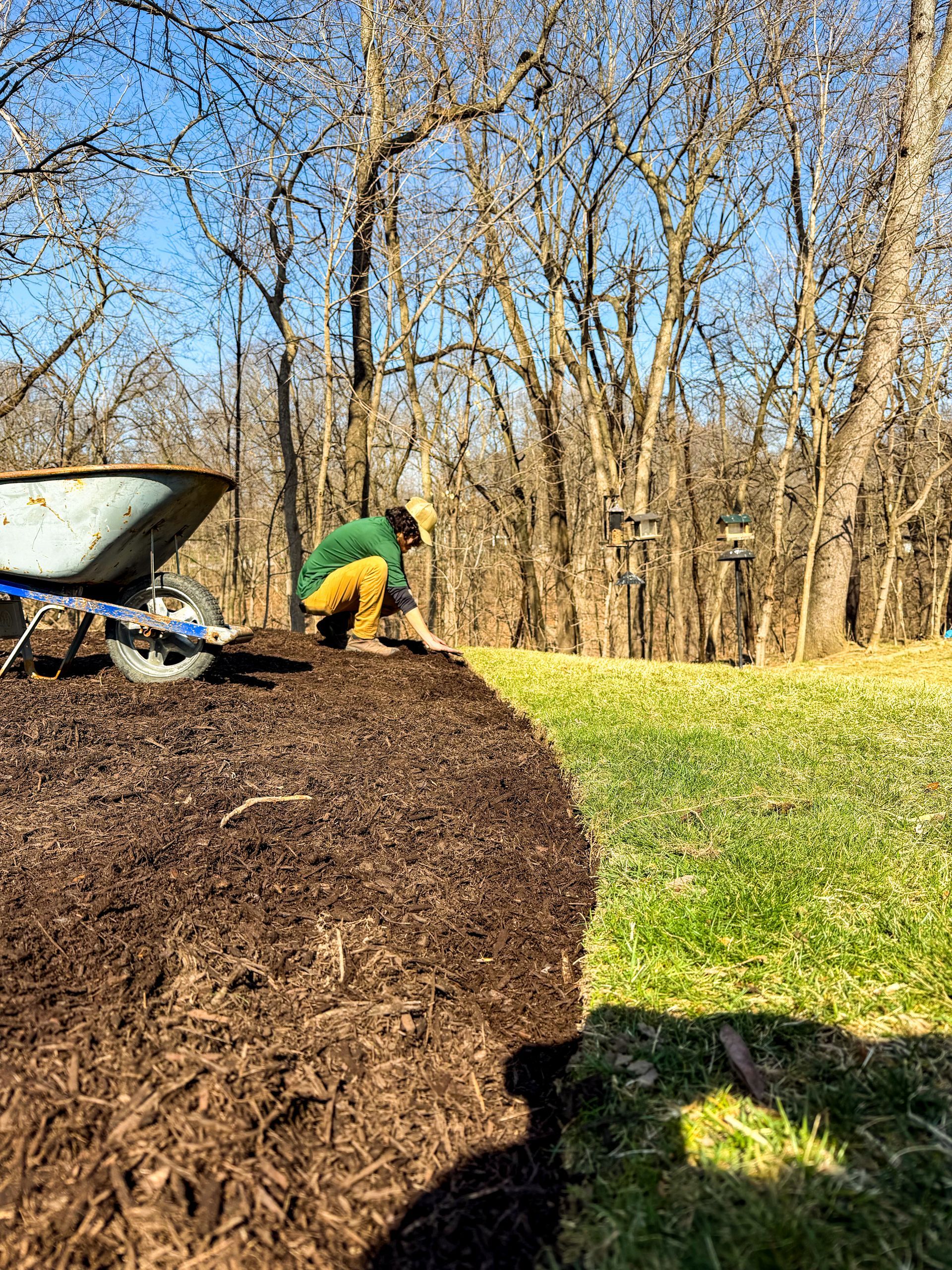 Person in green jacket and yellow pants working beside a large mulch pile in a sunny wooded yard.