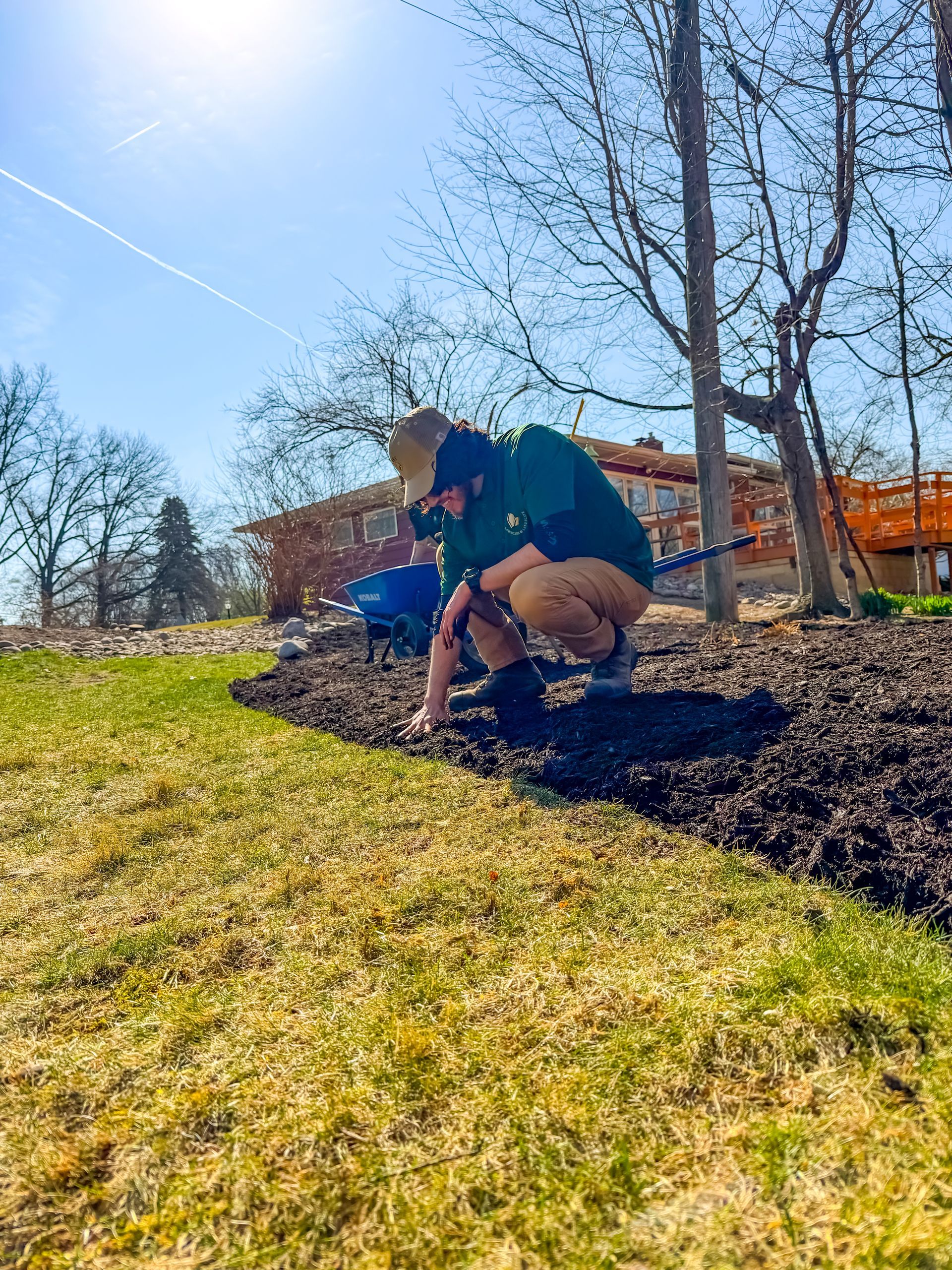 Person crouching on a sunny grassy slope beside a leafless tree and orange building, looking at a blue object