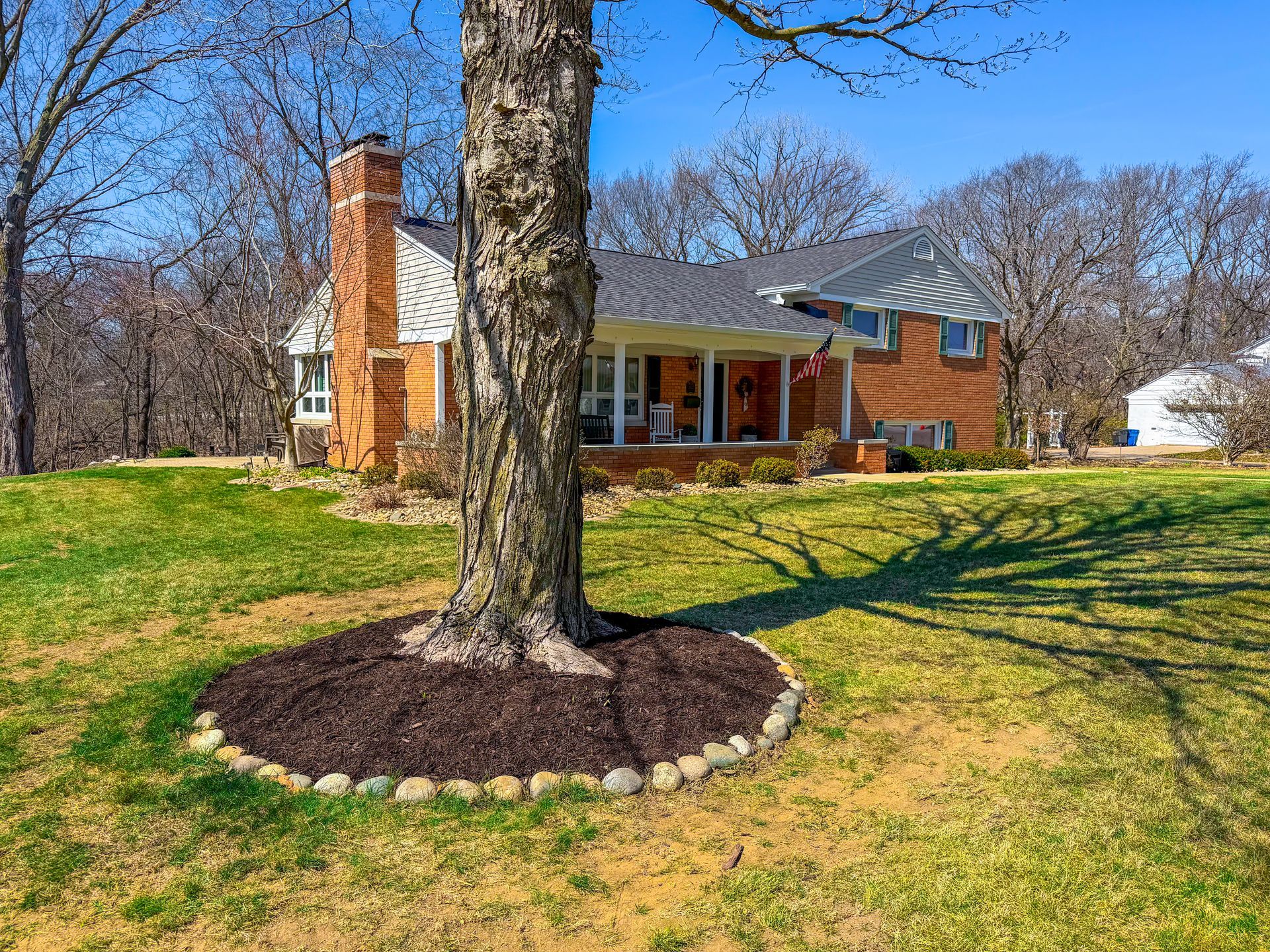 Brick house with a front porch, large tree, and grassy yard under a clear blue sky