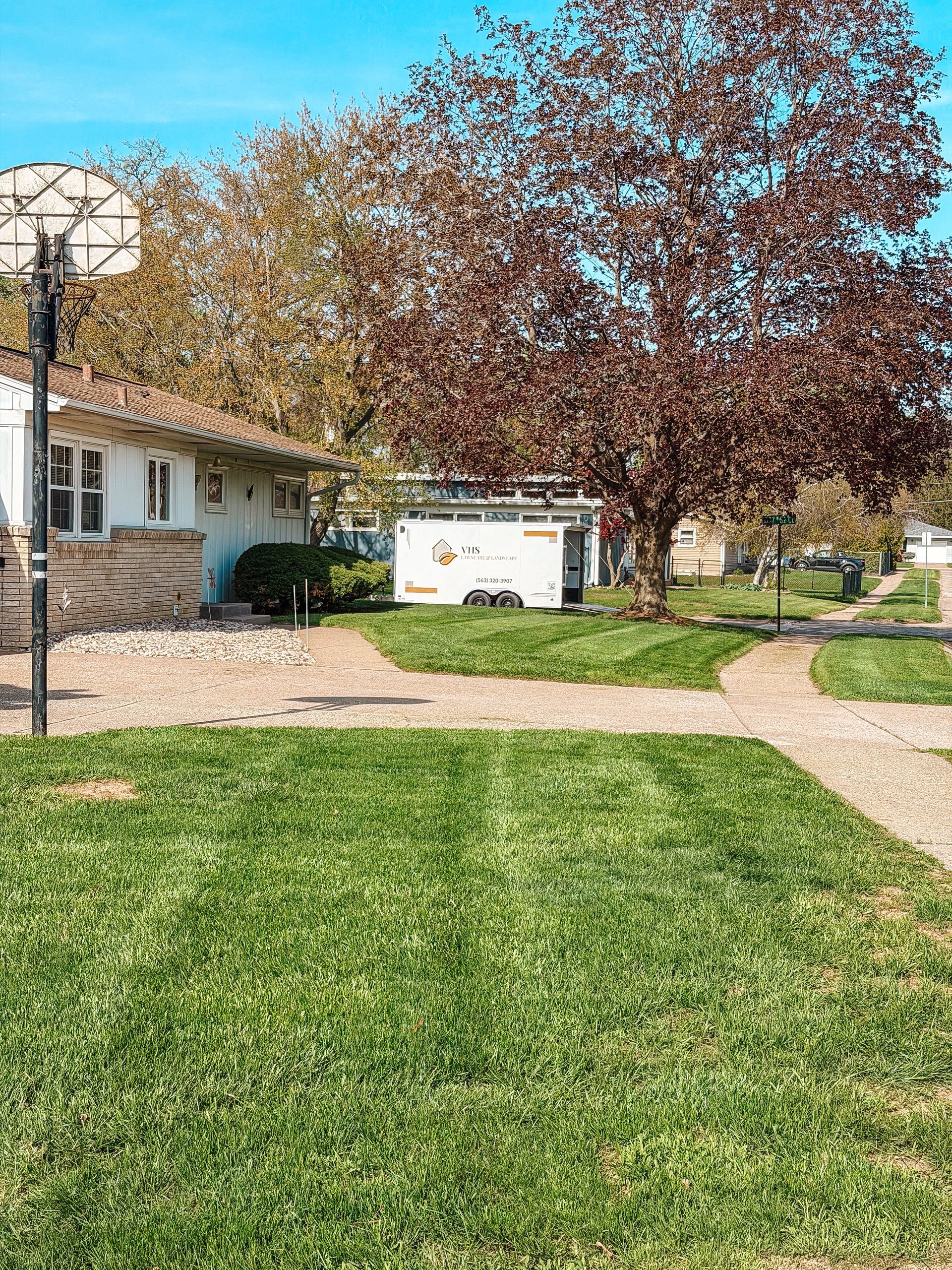 Suburban yard with green lawn, brick house, large tree, and a white utility box near a walkway.