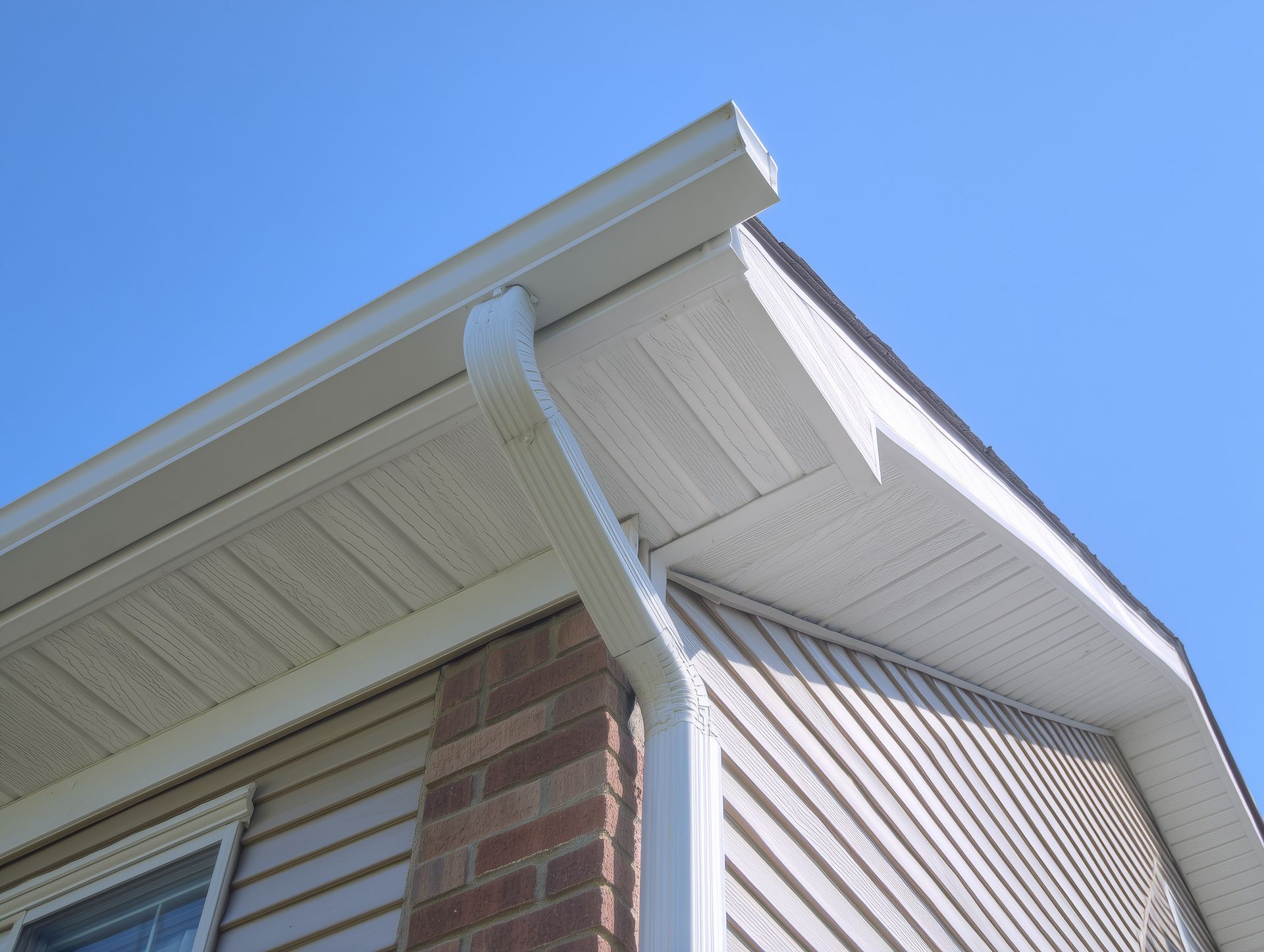 Système de gouttière blanche sur un coin de maison en briques et bardage beige, sur fond de ciel bleu.