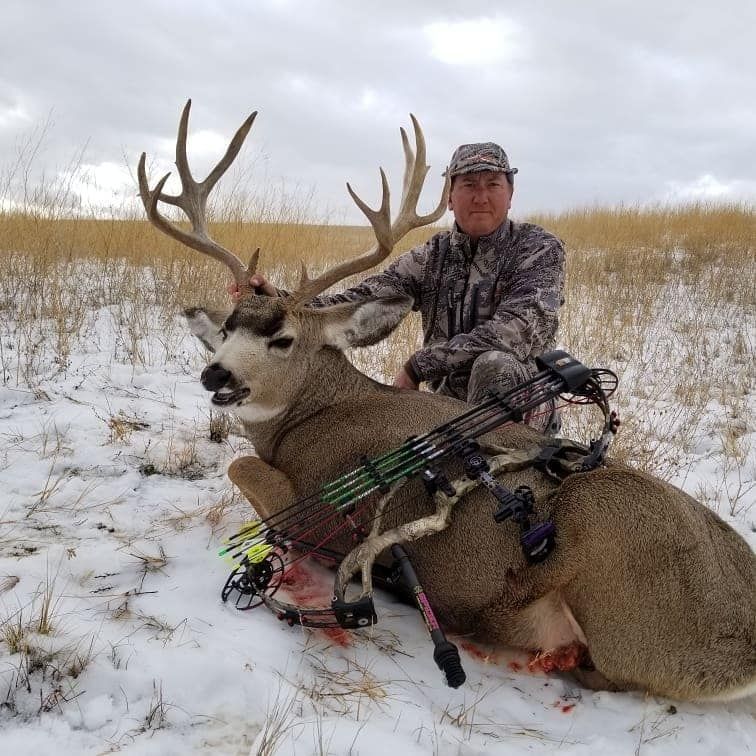 Hunter poses with a large deer on snow-covered ground, wearing camouflage and holding a crossbow.
