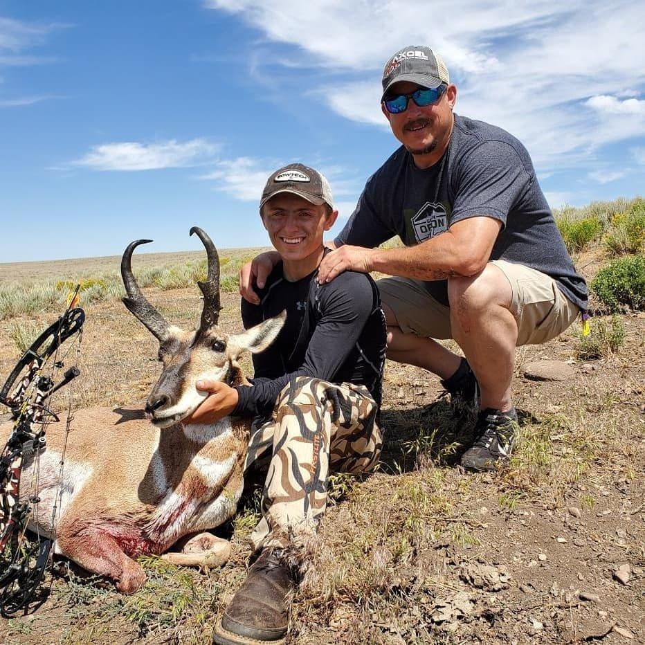 Two hunters kneel beside a harvested antelope in a rocky desert landscape under blue sky