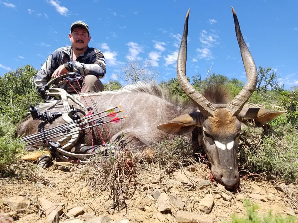 Hunter kneels beside a dead antelope with large horns in a dry, rocky field under a blue sky.