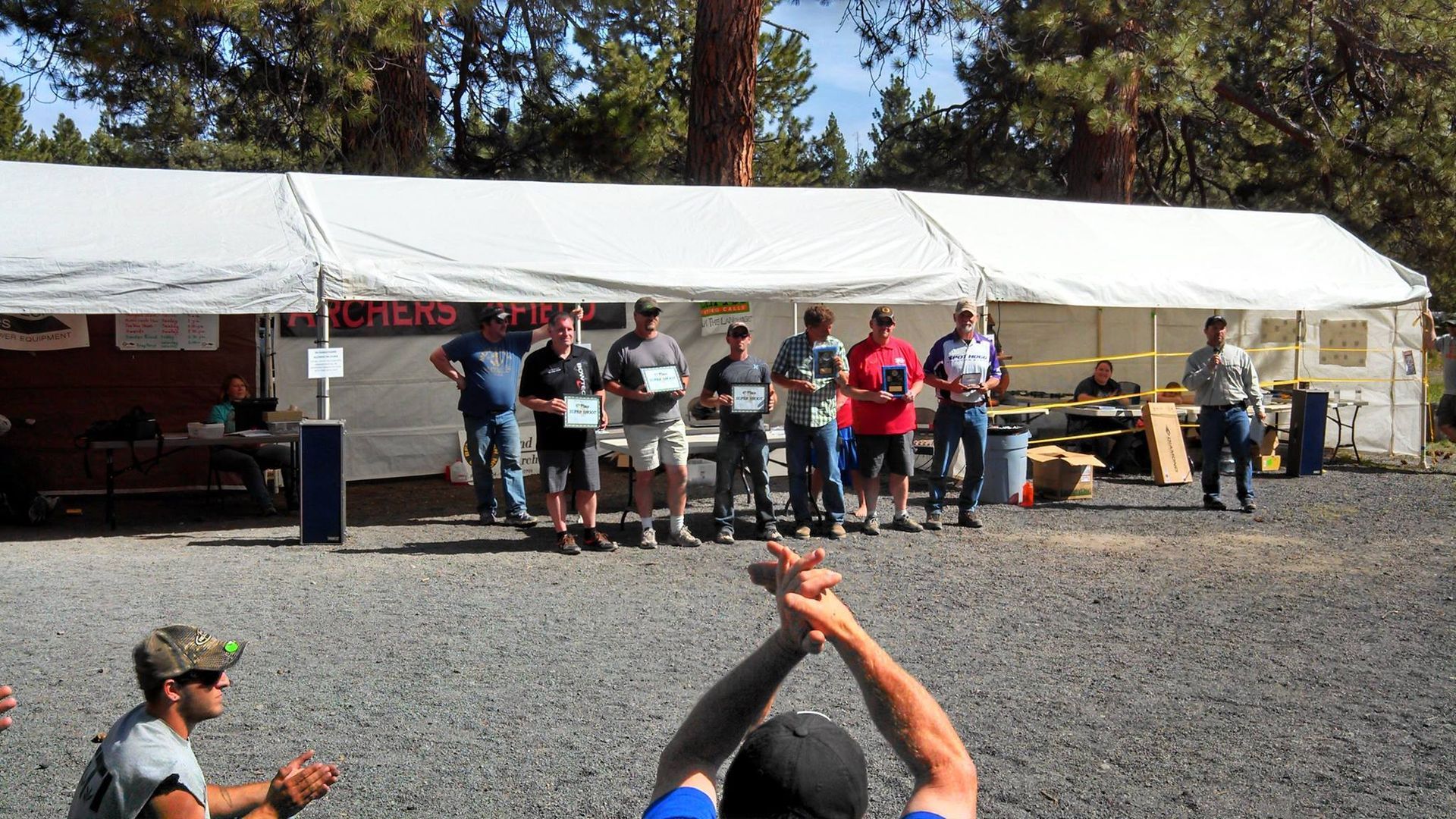 People standing under a white canopy at an outdoor event, with a gravel lot and trees behind them.