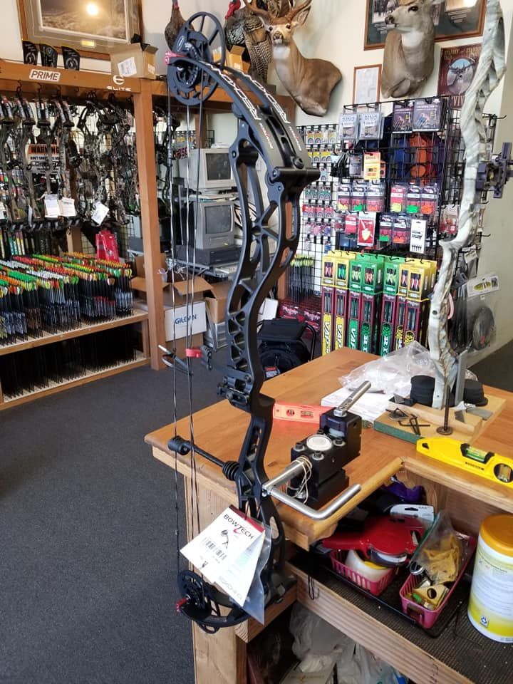 An archery shop display with compound bows, accessories, and gear on shelves and tables.