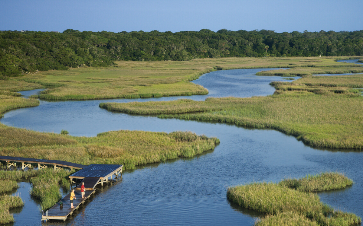 National Estuary Program logo in the bottom-left corner of a photo showing two individuals fishing off a dock in a marsh.