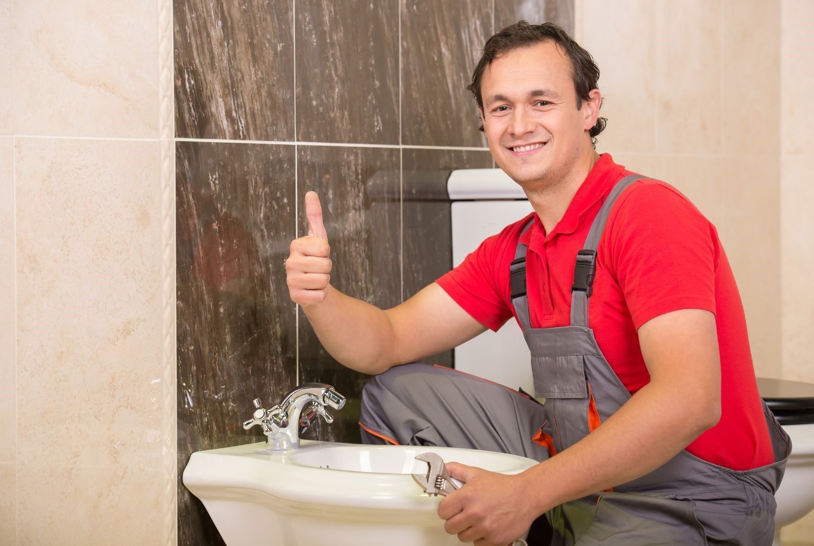 A plumber is kneeling on a bidet in a bathroom and giving a thumbs up.