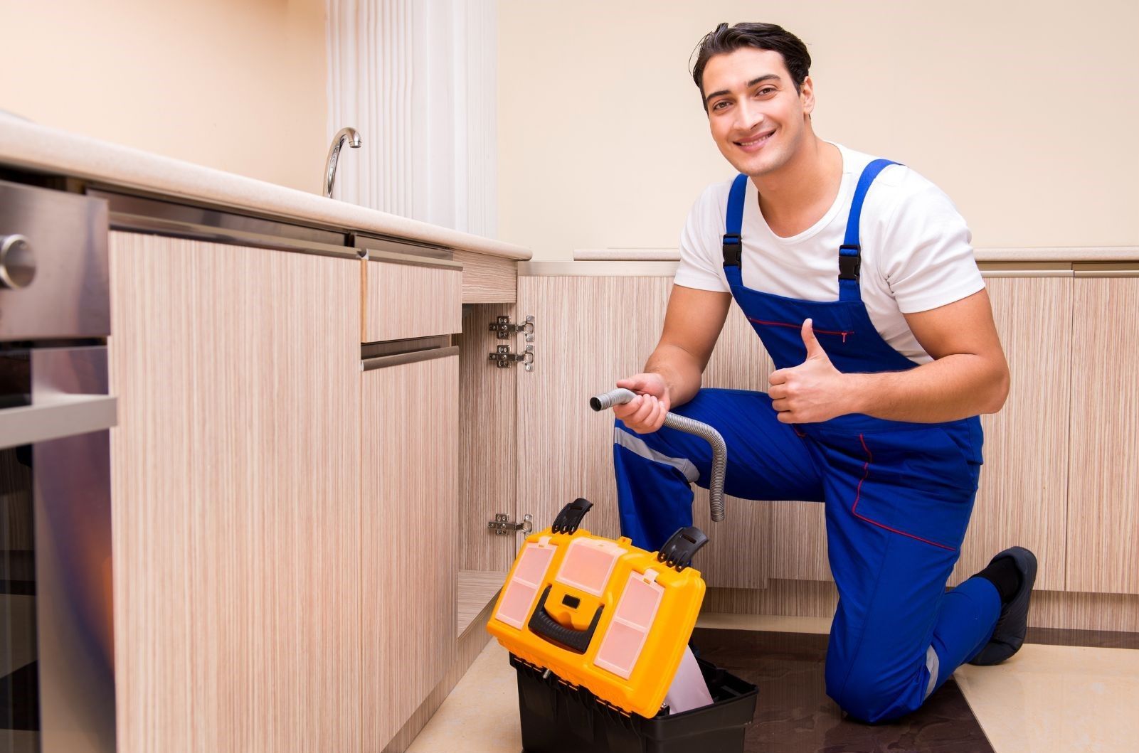 A plumber is fixing a sink in a kitchen and giving a thumbs up.