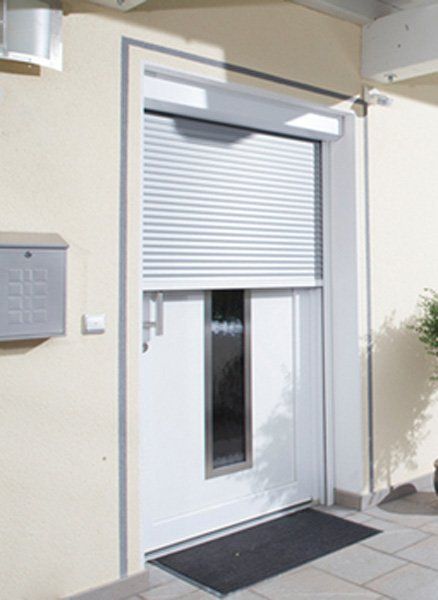 Ingresso dell'edificio con una serranda avvolgibile bianca parzialmente chiusa sopra una porta e una finestra stretta.