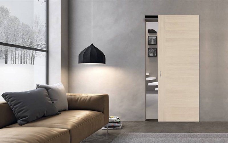 Soggiorno moderno con divano color cuoio, lampada a sospensione nera, ampia finestra e porta scorrevole in legno chiaro.