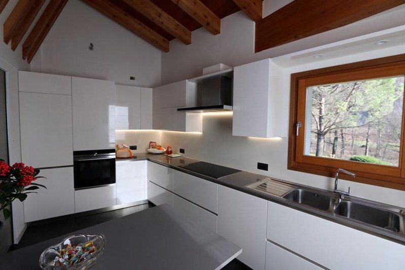 Cucina moderna bianca con soffitto con travi a vista, ripiani neri e un'ampia finestra con vista sugli alberi.