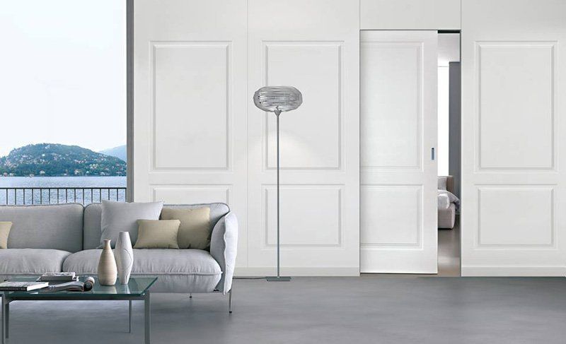 Luminoso soggiorno con divano grigio, tavolo rotondo, ventilatore a piantana e porte scorrevoli bianche che si aprono su una vista sull'acqua.