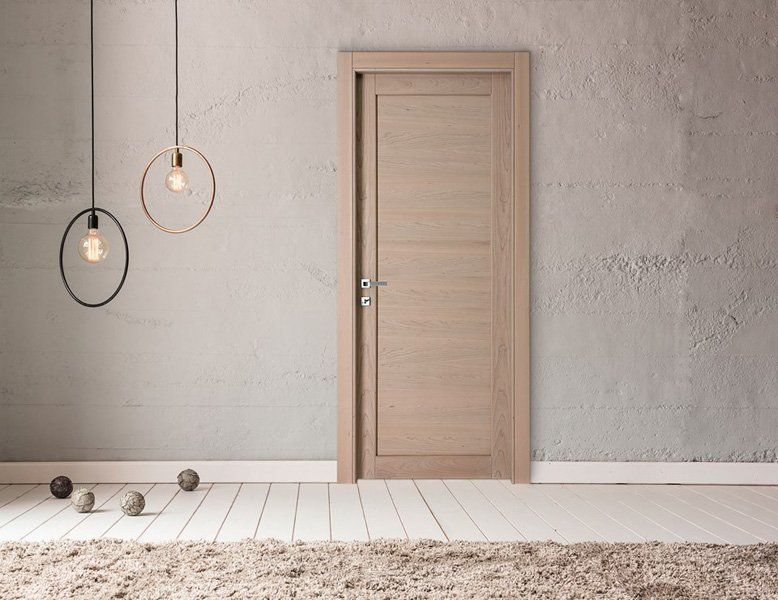 Interno minimalista con porta in legno chiaro chiusa, lampade a sospensione e parete bianca con finitura texturizzata.