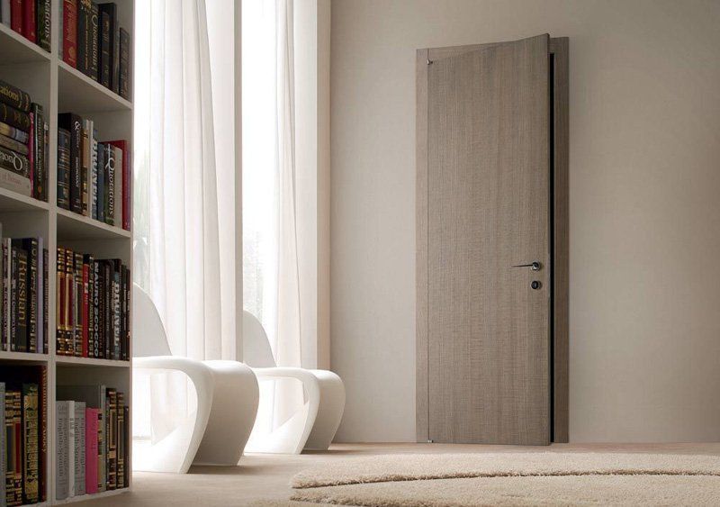 Stanza minimalista con libreria, sedie bianche e una porta grigia chiusa accanto a tende trasparenti.