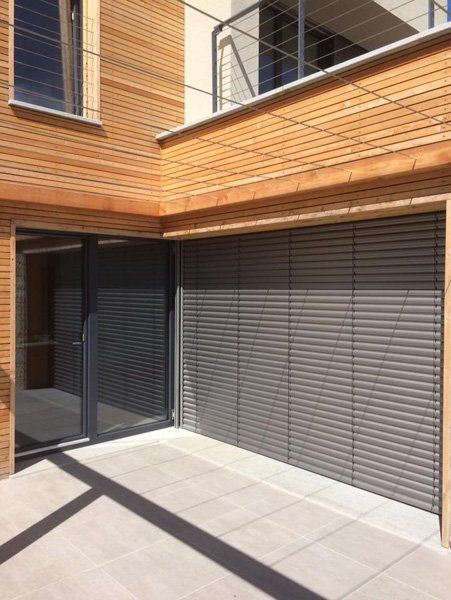 Cortile moderno con rivestimento in legno, persiane grigie e porte scorrevoli in vetro sotto un balcone.