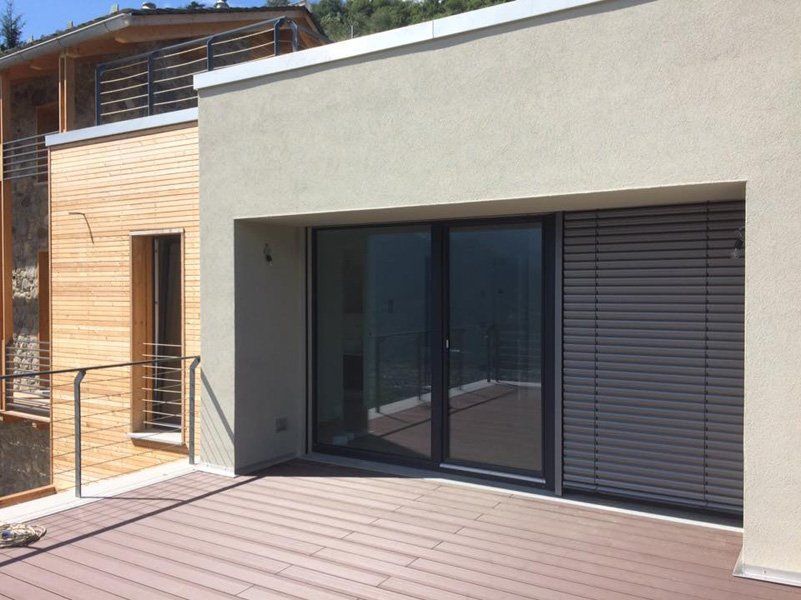 Patio di una casa moderna con porte scorrevoli in vetro, facciata beige e ringhiera in metallo su una terrazza in legno.