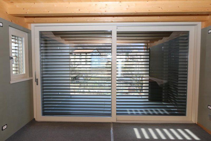 Camera moderna con ampie porte scorrevoli in vetro coperte da tende orizzontali e giochi di luce sul pavimento.