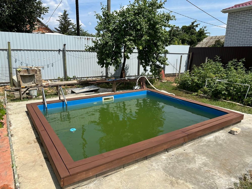 Small backyard pool with green water, wooden deck edging, and patio chairs under a sunny sky