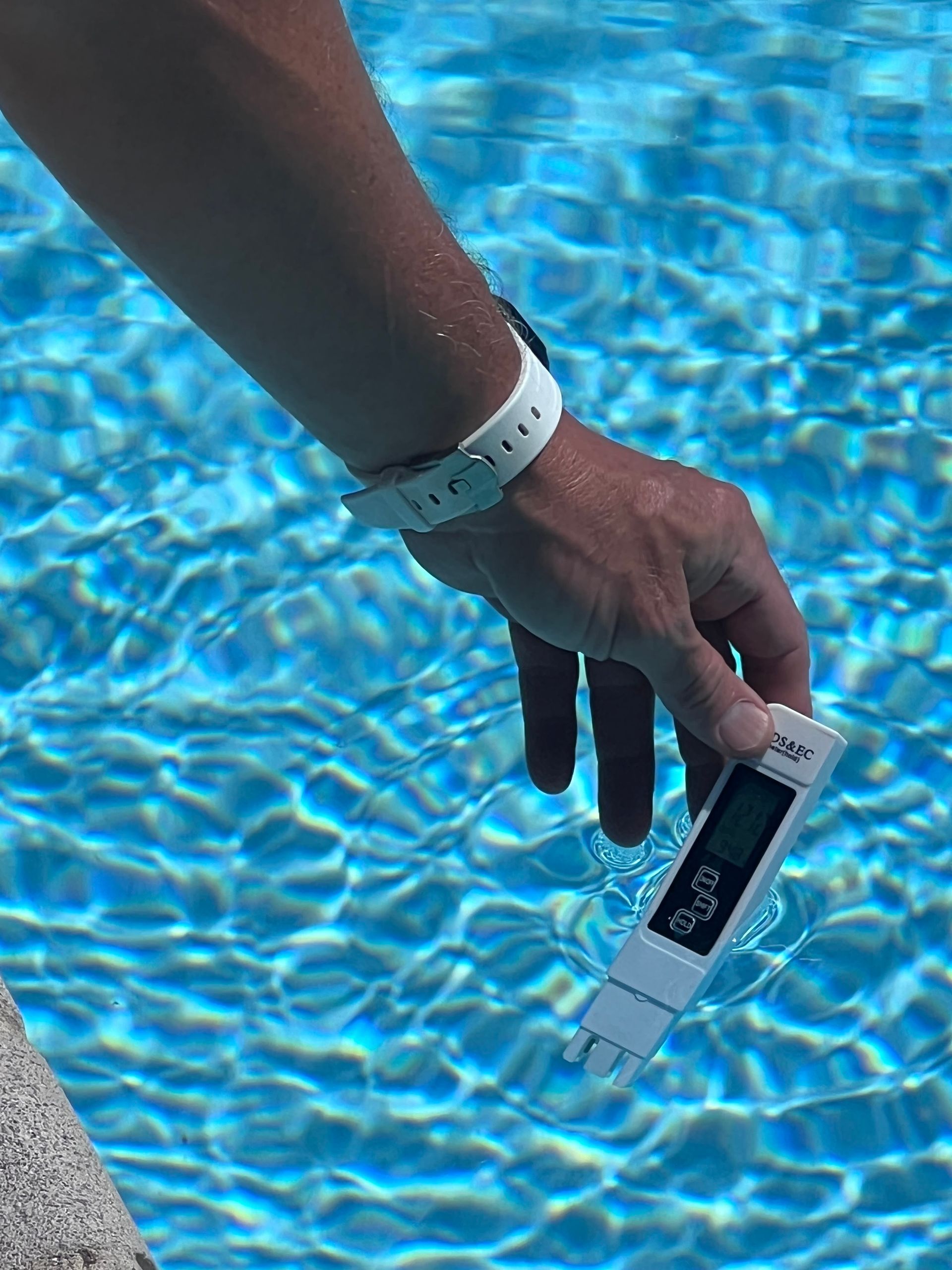Hand holding a digital pool water tester over a blue swimming pool