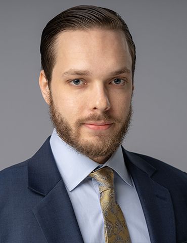 Professional headshot of a man in a navy suit, light blue shirt, and gold tie on a gray background