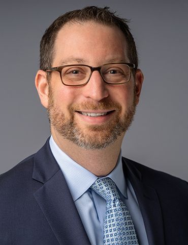Smiling man in glasses wearing a navy suit, light blue shirt, and patterned tie against a gray background