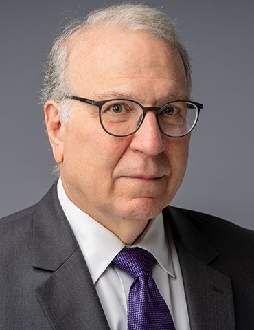 Portrait of a man in glasses and a gray suit with a purple tie, against a plain gray background