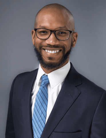 Smiling person in glasses and navy suit with blue striped tie against gray background