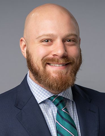 Smiling bald man with a red beard in a dark suit, white shirt, and teal striped tie against a gray background