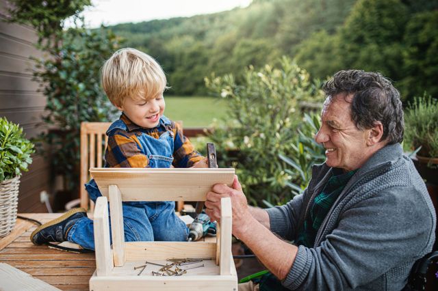 Man and child smiling while building a wooden chair outdoors in a garden