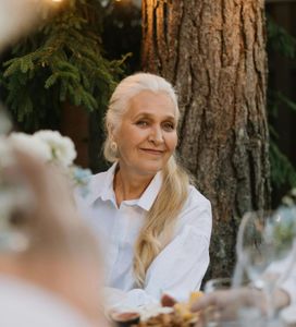 Woman in a white shirt seated outdoors beside a tree, smiling softly at a table setting.