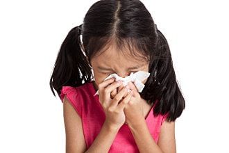 Girl in a pink shirt blowing her nose with a tissue, eyes closed.