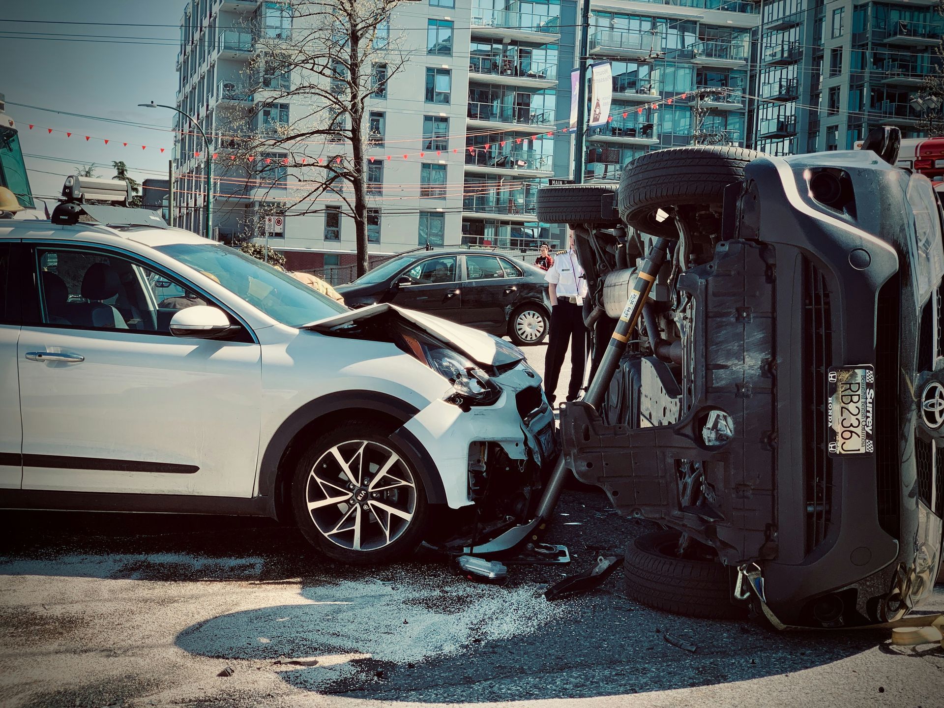Car accident scene: White SUV damaged, black SUV overturned, bystanders, urban setting.