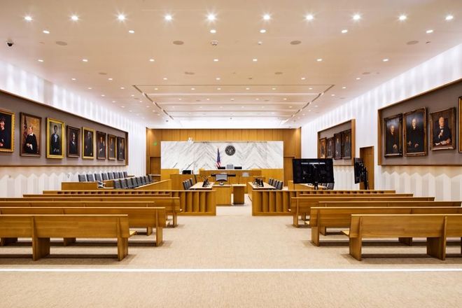 Empty courtroom with wooden benches, framed portraits, and a judge’s bench at the front