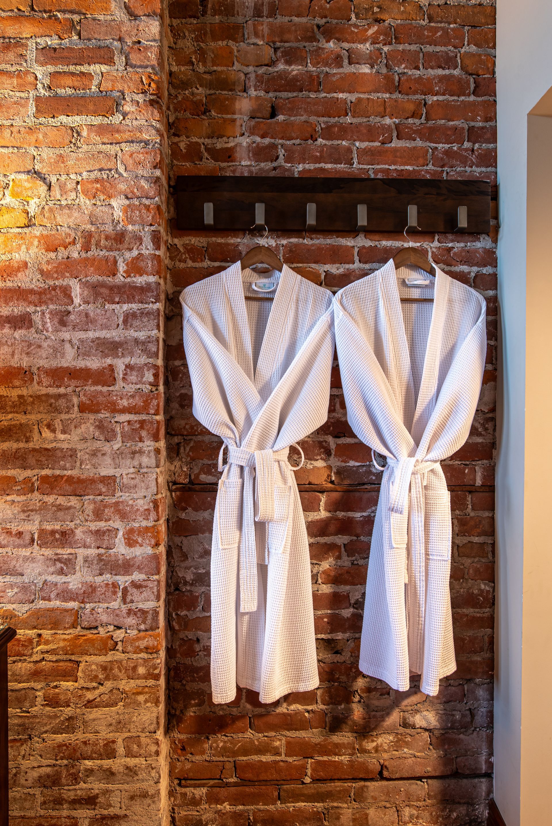 Two white bathrobes are hanging on a brick wall.