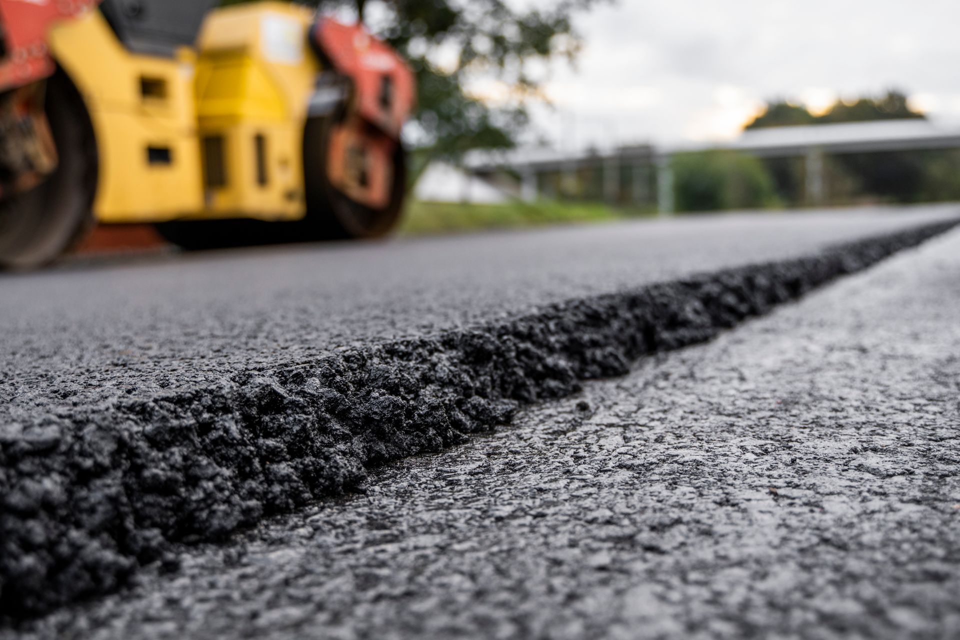 Low-angle view of freshly laid asphalt edge with a road roller in the background.