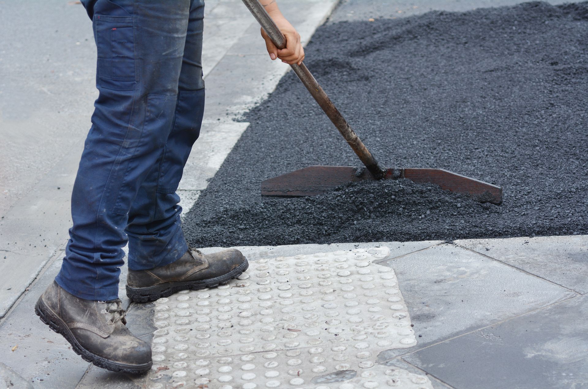 Worker spreading and leveling fresh asphalt near a sidewalk curb during pavement repair work.