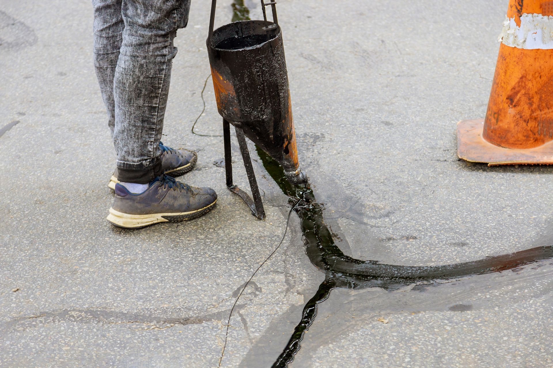 Person pouring black sealant into a crack in asphalt pavement with an orange traffic cone nearby.