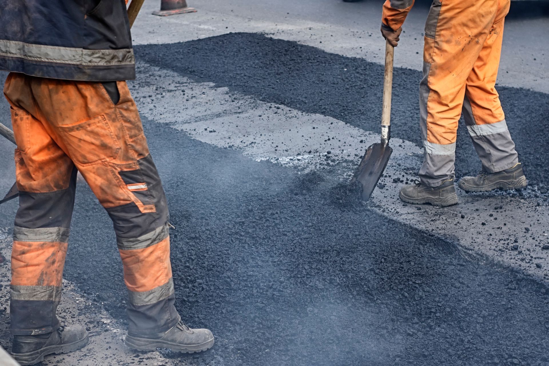 Two workers in orange workwear are shoveling asphalt onto a road.