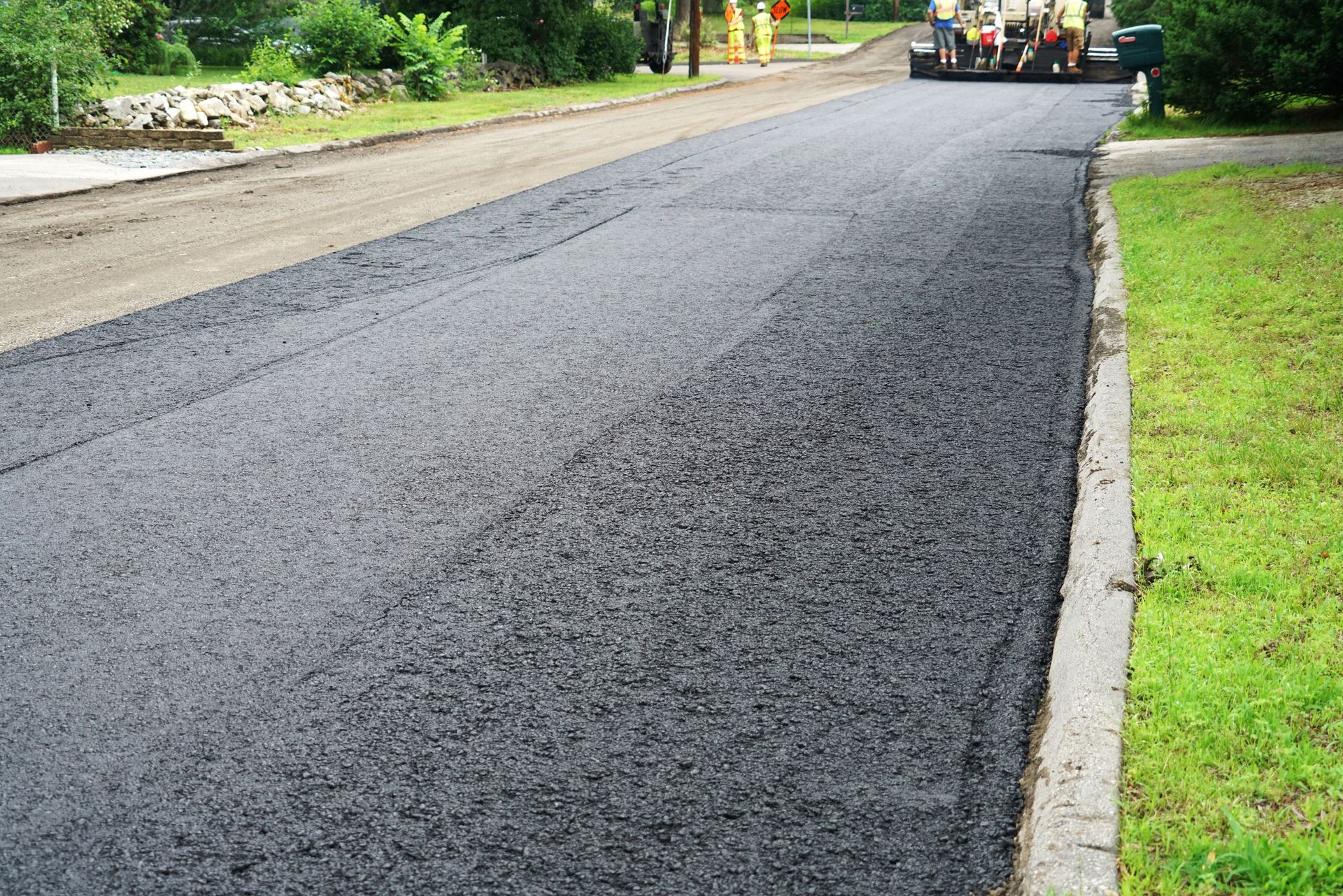 Freshly paved asphalt road; asphalt roller in background; green grass and curb on right.