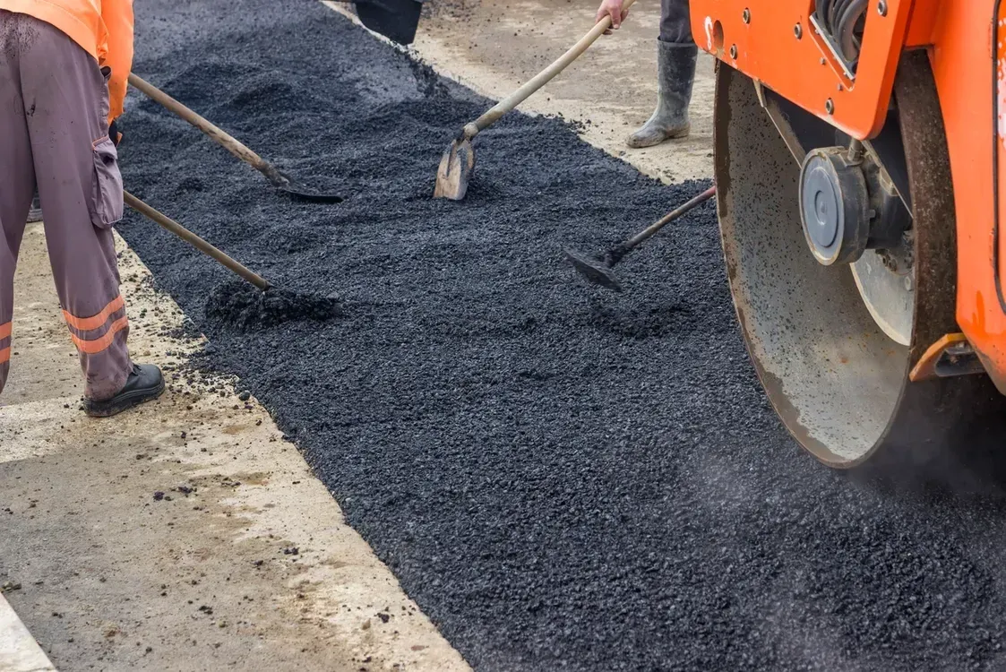 Road construction: workers laying asphalt with rakes, orange roller in the background.